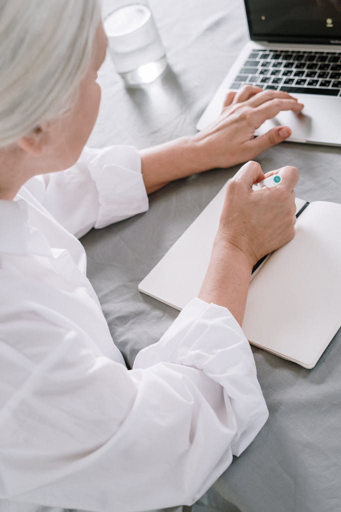 Elderly woman takes notes while using a laptop at home.